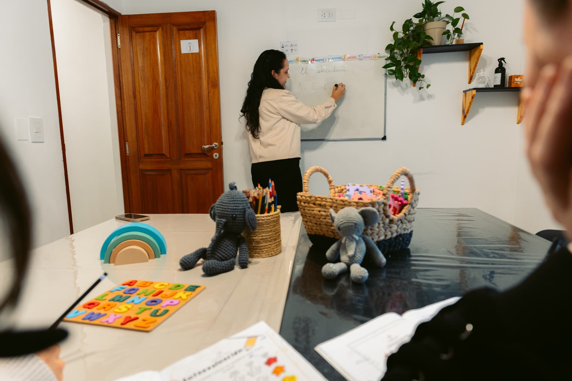 Una profesional de Centro ANPI escribe el abecedario en el pizarrón del aula de apoyo escolar, con material didáctico sobre las mesas del espacio.