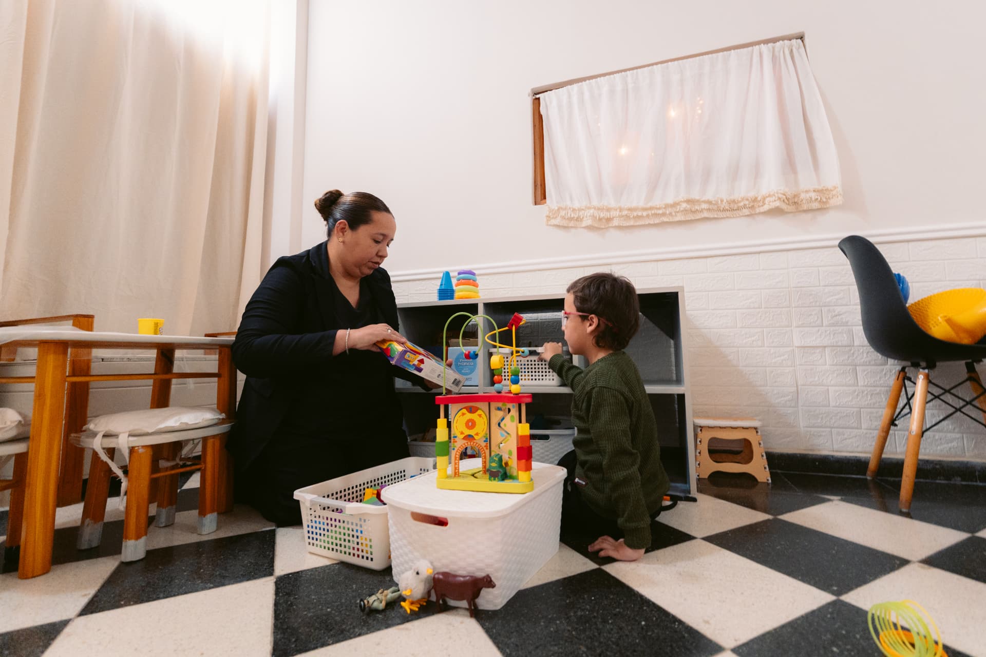 Una profesional de Centro ANPI acompaña a un niño que juega con piezas y bloques en el piso del espacio de talleres, entre estantes con materiales organizados y una mesa infantil de madera.