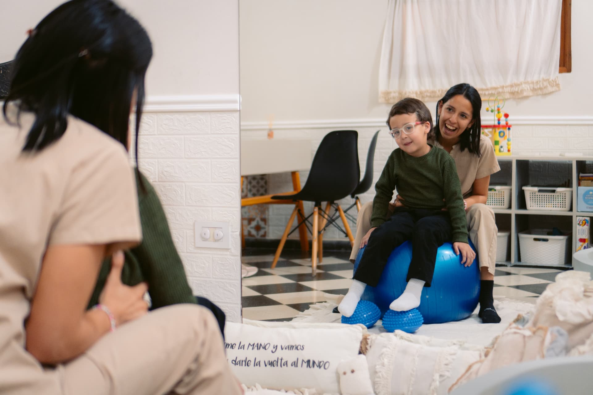 Un niño con lentes se sienta sobre una pelota de pilates azul mientras una profesional de kinesiología de Centro ANPI lo sostiene, frente al espejo de la sala del Centro.