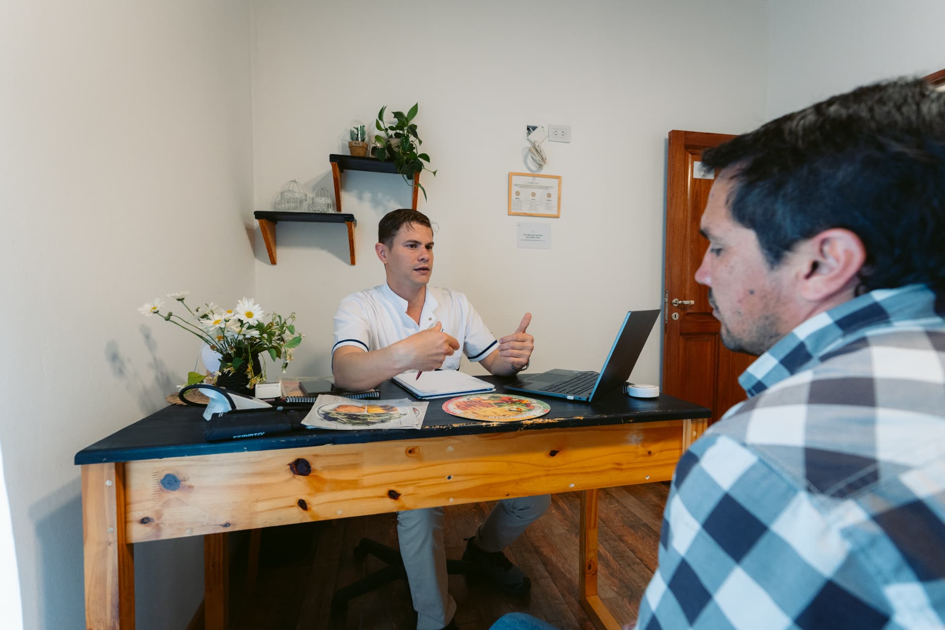 Hernán Vecchia, Lic. en Nutrición, conversa con una persona sentada frente a su escritorio en el consultorio de Centro ANPI, con láminas de alimentación sobre la mesa.