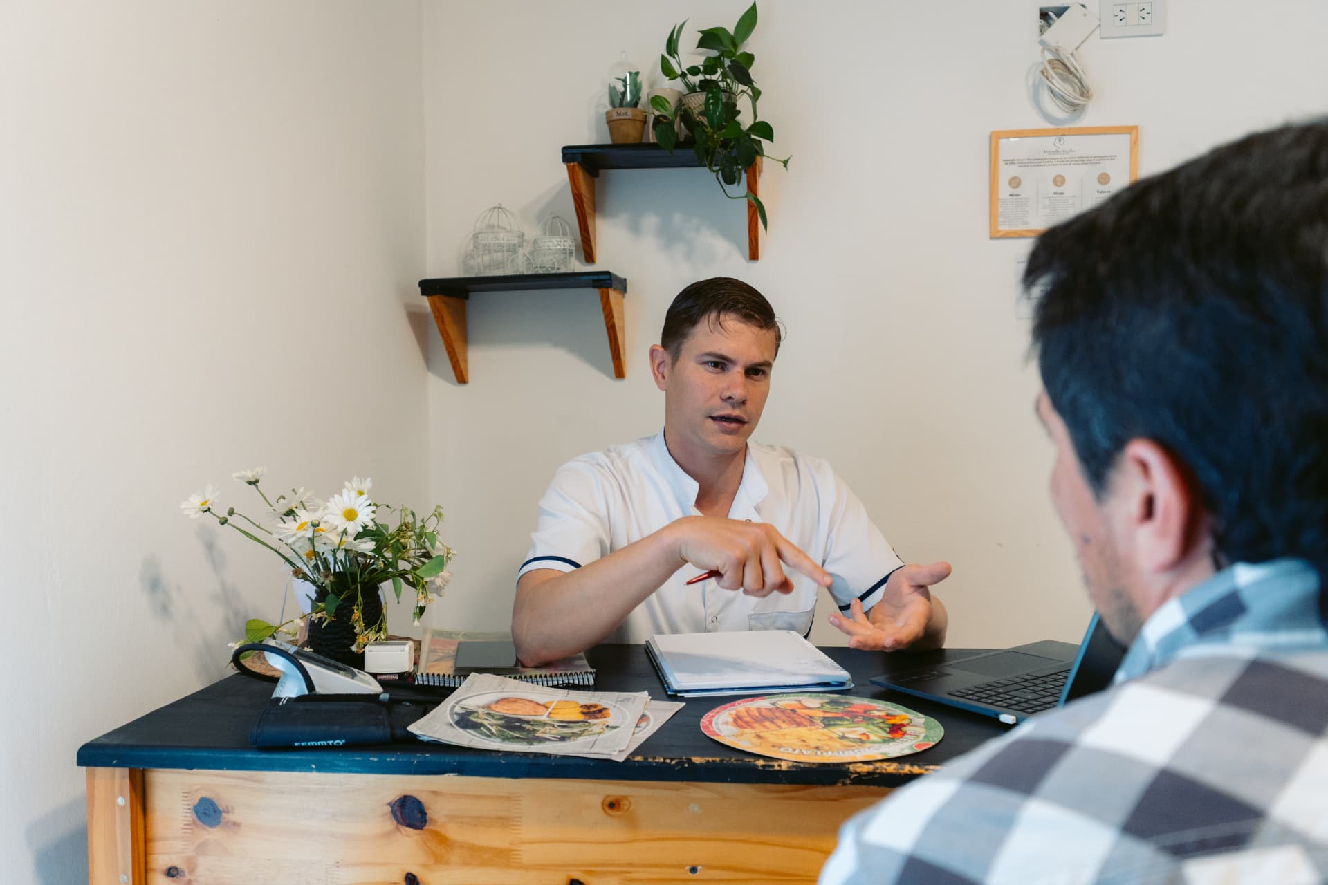 Hernán Vecchia, del servicio de Nutrición de Centro ANPI, conversando con una persona sentada frente a su escritorio en el consultorio, con láminas sobre alimentación sobre la mesa.